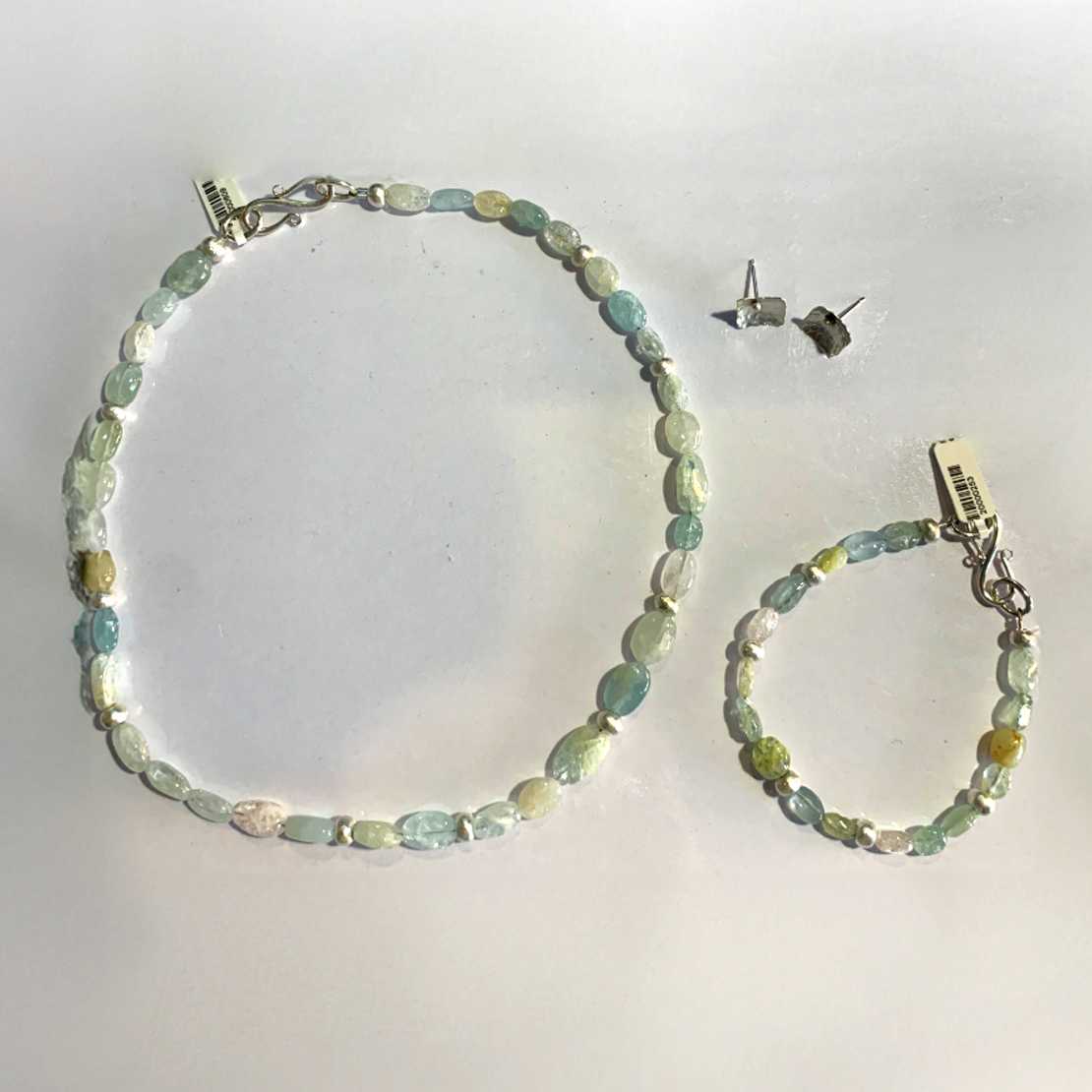 This image displays a delicate necklace made of oval-shaped fluorite beads in varying shades of green, blue, and light yellow with matching bracelet and two square silver studs. The beads are interspersed with small silver spacer beads, adding contrast. The necklace is fastened with a simple silver hook-and-loop clasp. The translucent nature of the beads gives it an elegant and organic feel, reminiscent of sea glass or natural gemstones.