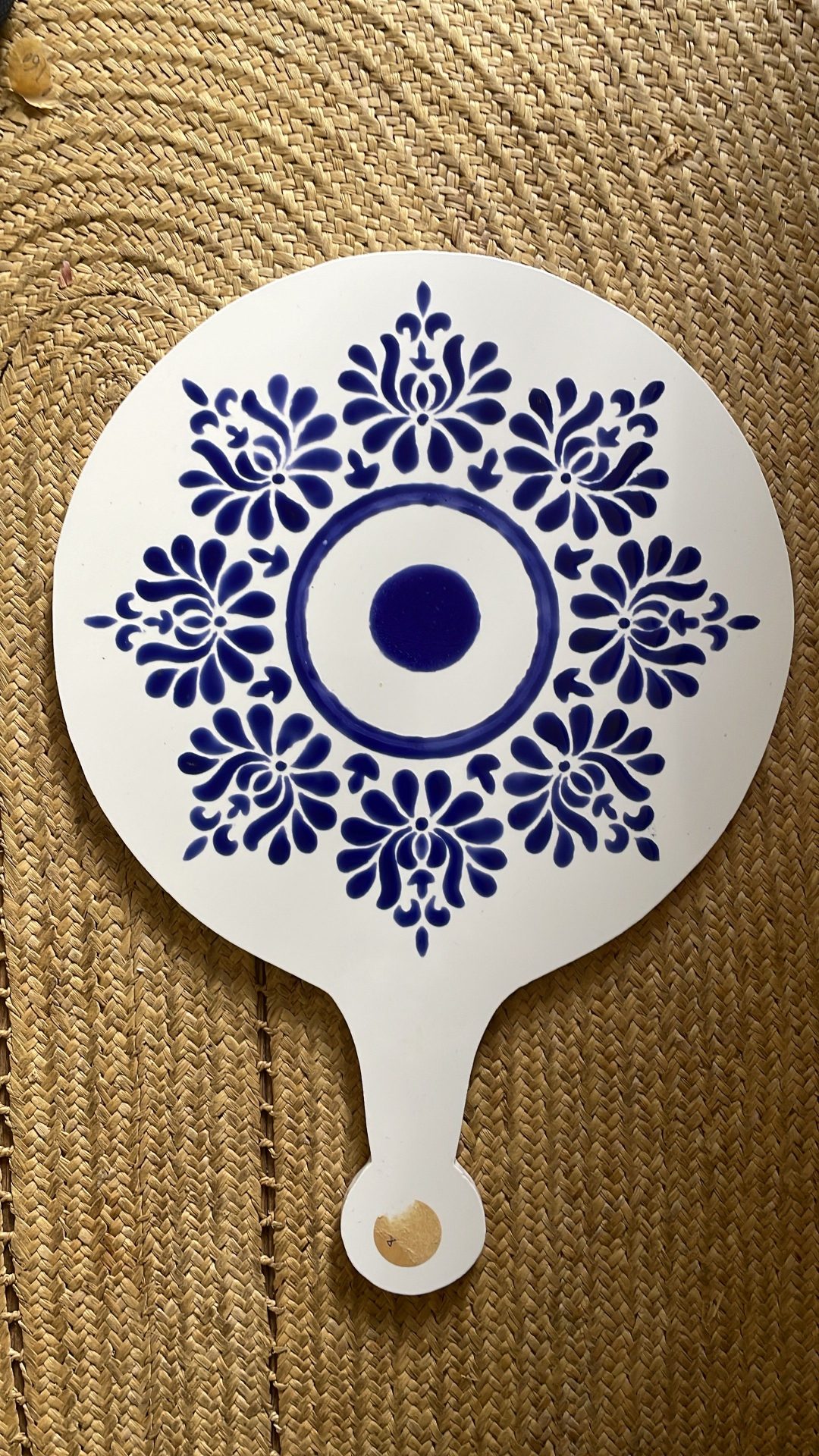 Blue and White Medallion Cheeseboard This is a white ceramic cheeseboard with a round top and small handle. It features an elegant blue and white design reminiscent of traditional porcelain patterns. The central motif has concentric circles with a solid blue dot in the middle, surrounded by ornate floral patterns arranged in a symmetrical medallion design. The board is displayed on a woven natural fiber mat or placemat with a braided texture.