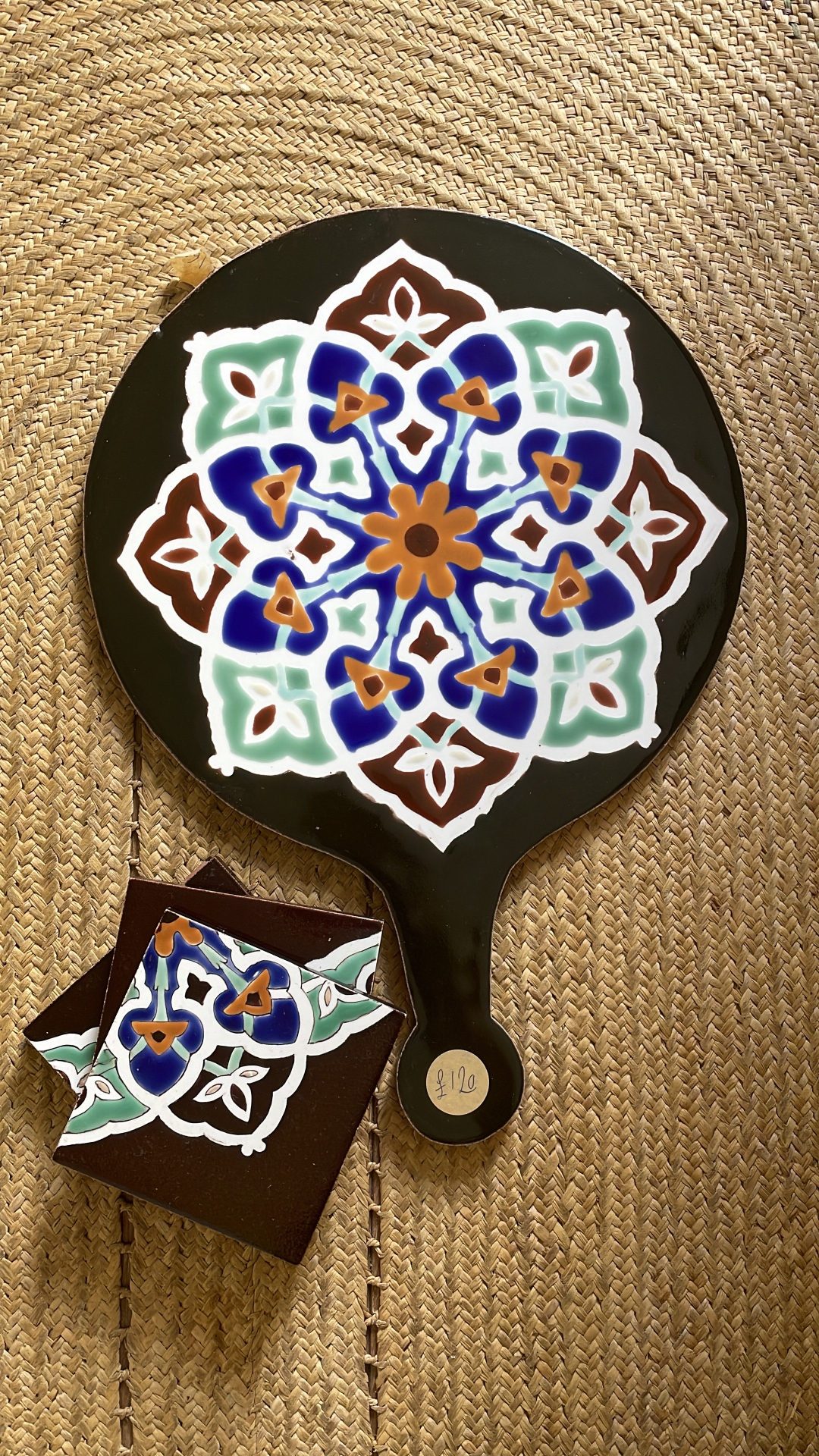 This ceramic cheeseboard has a black background with a colorful mandala-like pattern. The intricate design features a symmetrical arrangement of blue, mint green, white, and orange/brown elements forming a floral star pattern. The handle is visible at the bottom, and there appears to be a small matching coaster or sample piece with a similar pattern placed beside it. Like the first image, it's displayed on the same type of woven natural fiber mat.