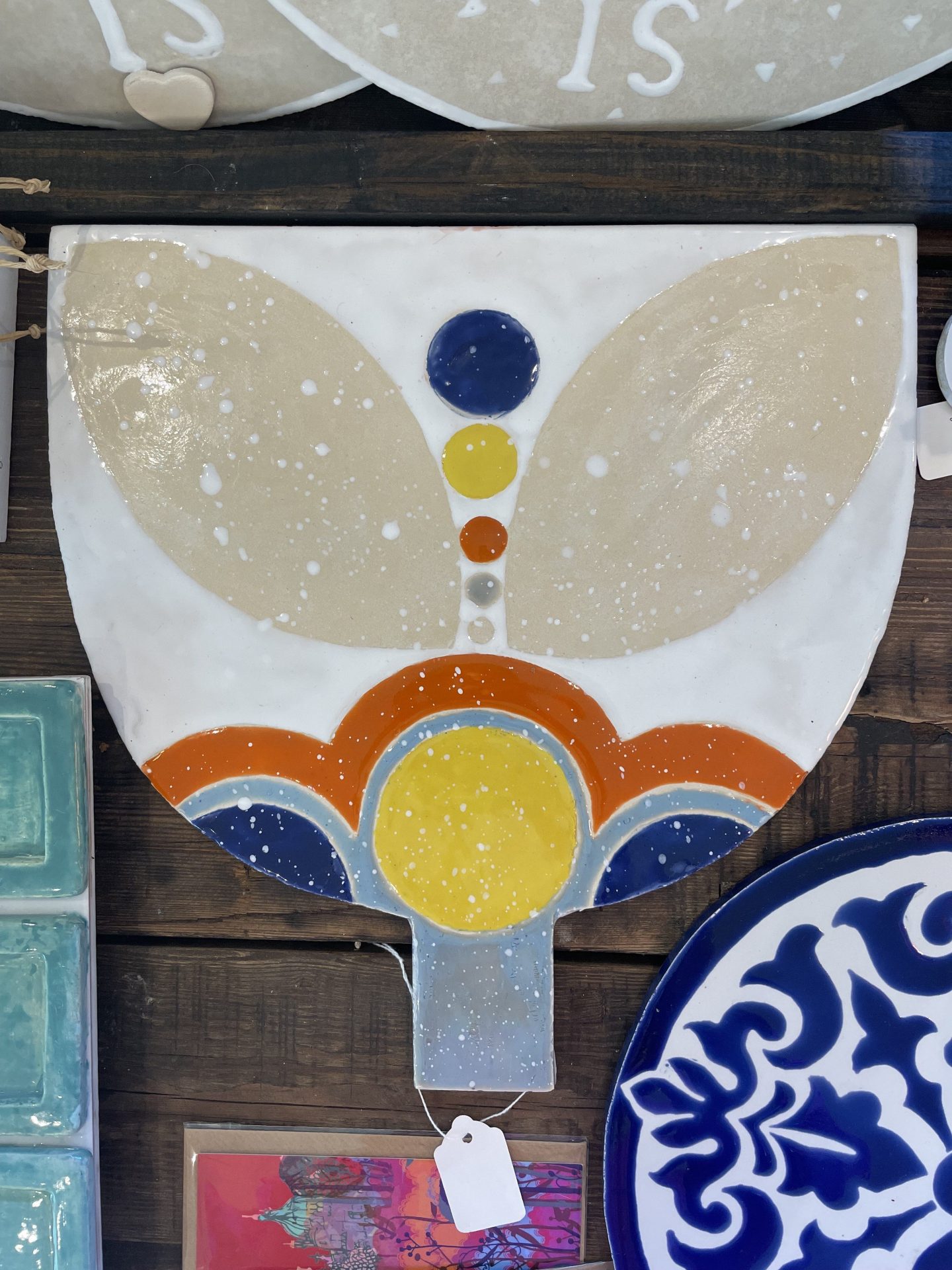 Floral Cheeseboard This artistic ceramic serving board has a white background with a stylized butterfly or angel-like design. The "wings" are a beige/cream color with white speckles, while the central "body" features a vertical line of colorful dots (blue, yellow, orange) and a decorative base with curved elements in blue, orange, and yellow. It has a small handle at the bottom and appears to be displayed on a wooden surface alongside other ceramic pieces, including what looks like turquoise tiles and a blue and white plate with a floral pattern.