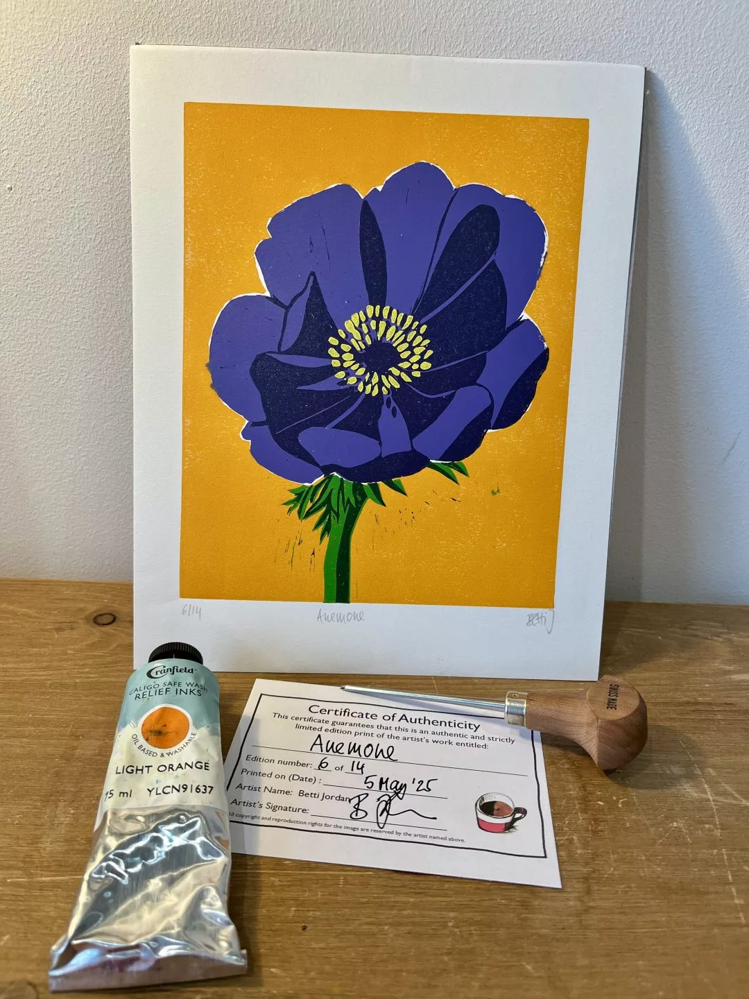 A framed print of a large purple anemone flower with layered petals and a yellow center is propped upright against a wall on a wooden surface. The background of the print is a solid bright orange. Below the print lies a certificate of authenticity, a metal tube of light‑orange printing ink, and a wooden‑handled roller tool.