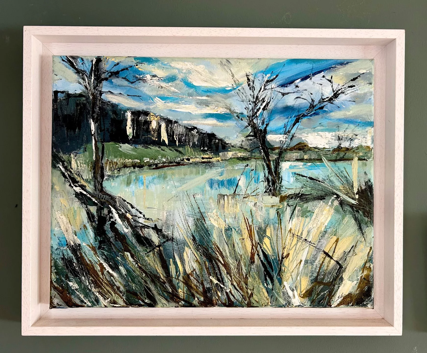 A framed landscape painting showing a riverside or lakeside scene. In the foreground, tall grasses and reeds in shades of beige, green, and brown stretch upward with bold, textured brushstrokes. Behind them, calm reflective water occupies the center of the painting, capturing patches of light blue and soft yellow highlights. On either side of the water, two leafless trees with dark, expressive branches stand partly submerged. A long, dark hillside runs across the left side of the background, contrasting with a bright sky filled with streaks of blue, white, and grey cloud patterns. The painting is framed in a light wooden frame and is shown hanging against a muted green wall.
