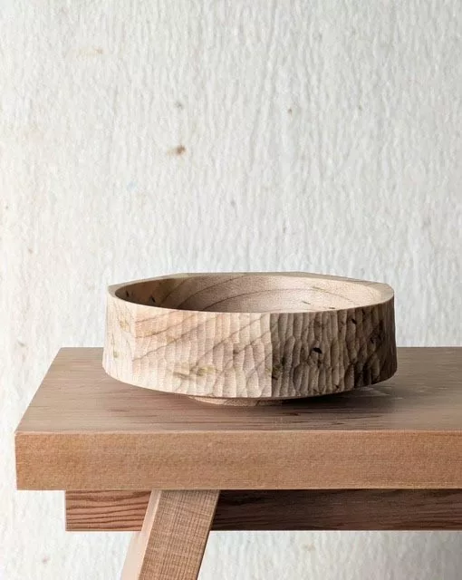 A handcrafted wooden bowl sits on a wooden surface. The bowl has a rough, textured exterior with visible carved grooves and tool marks, giving it a rustic, handmade feel. Its interior is smooth and polished, showing the natural grain of the wood. Behind it is a plain, light‑coloured wall that keeps the focus on the bowl’s craftsmanship and organic texture.