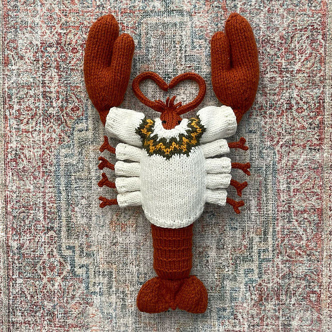 A hand‑knitted lobster plush lying on a patterned rug. The lobster has rust‑red claws, legs, and tail, with a cream‑coloured body and a decorative yellow, black, and brown motif around the top of the torso. The knitted texture is soft and detailed, giving the lobster a stylized, crafted appearance.