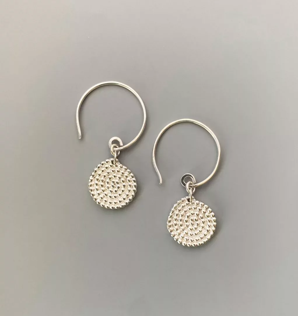 A pair of small silver drop earrings displayed on a smooth grey background. Each earring has a thin, circular hook and a round dangling charm. The charm is a flat disc covered in a raised, textured pattern made of tiny evenly spaced bumps arranged in concentric circles. The metal has a polished, reflective finish.