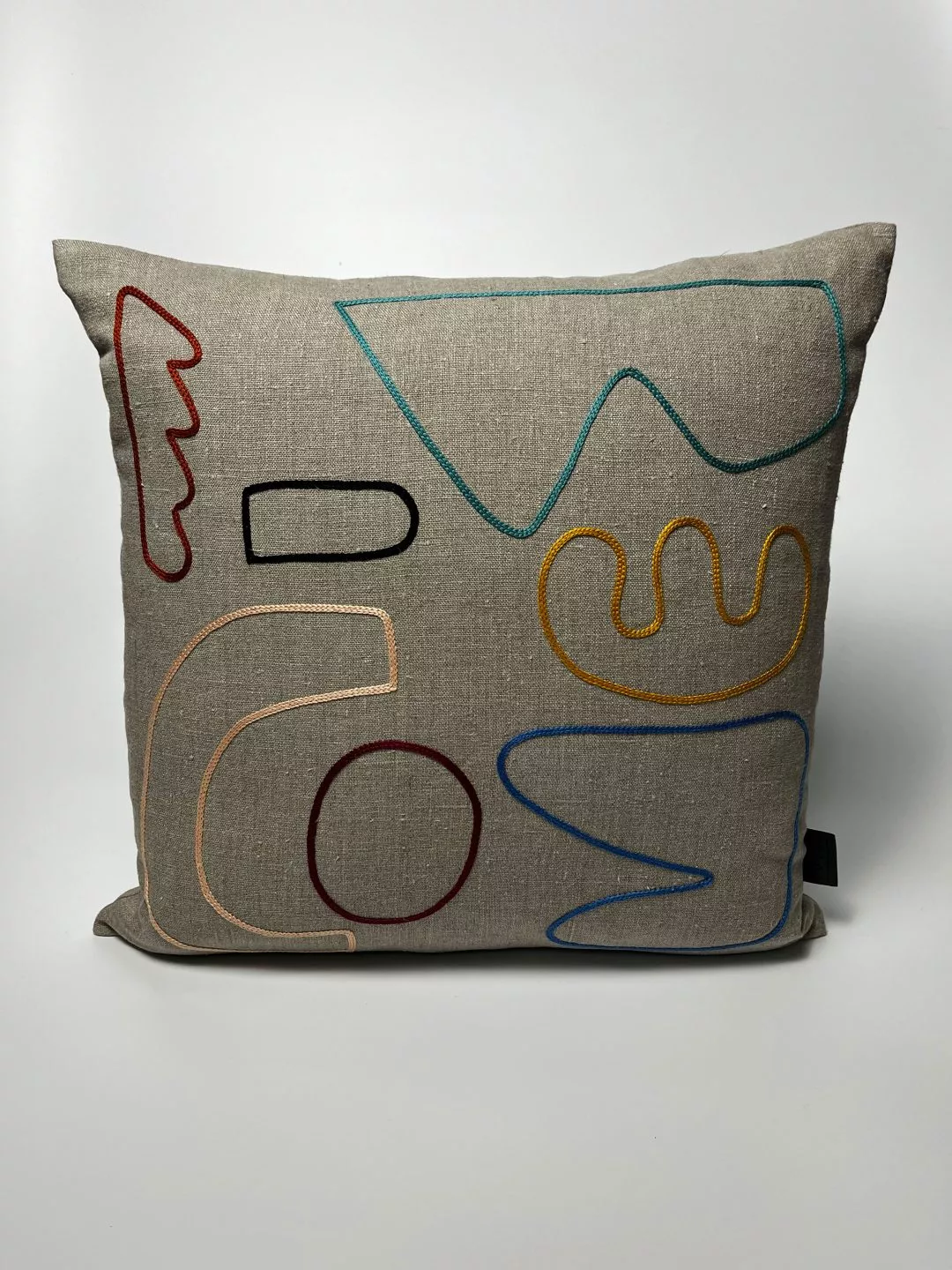 A square cushion with a light beige fabric cover shown against a plain white background. The front of the cushion features a series of abstract embroidered line shapes in various colors. These stitched lines include curved, looping, and angular forms in red, teal, yellow, black, peach, and blue. The shapes are arranged loosely across the surface without forming any recognizable image or text. The cushion appears soft and evenly filled, with slightly rounded corners and a small black fabric tag on the right edge.