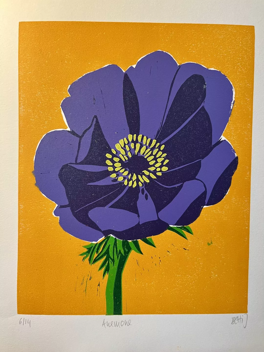 A close-up view of a print depicting a large stylised anemone flower. The flower has deep purple petals with darker purple shading and a cluster of small yellow shapes forming the center. The stem and leaves are bright green. The background is a flat, vivid orange.