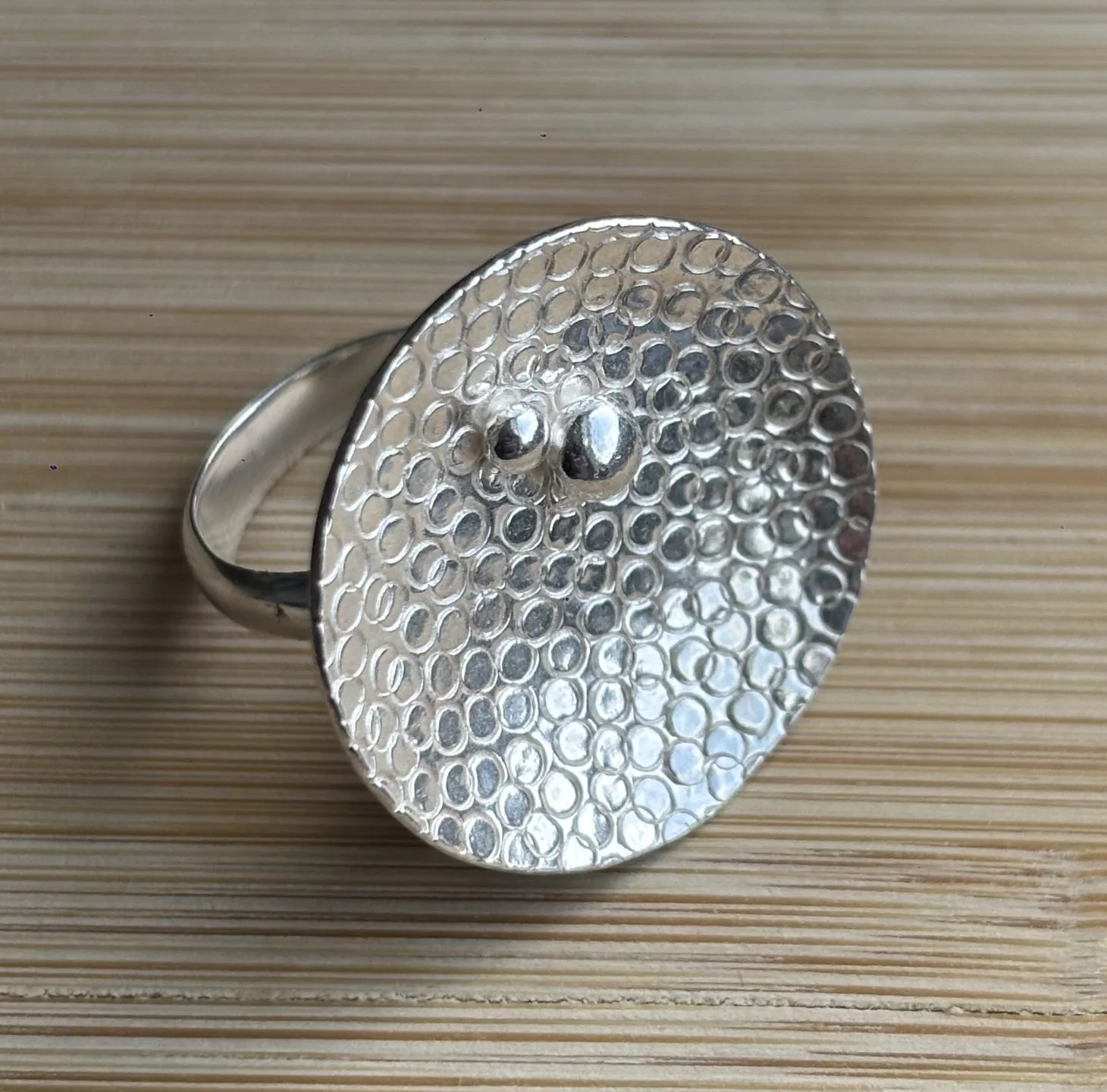 A silver ring displayed on a light wooden surface with fine horizontal grain lines. The ring features a large, flat, oval‑shaped top with a hammered texture made up of small, evenly spaced circular impressions. Near the center of the oval are two raised polished silver dots, one slightly larger than the other. The ring band is smooth and rounded, attached to the back of the oval top. The metal surface reflects light softly, highlighting the textured pattern.