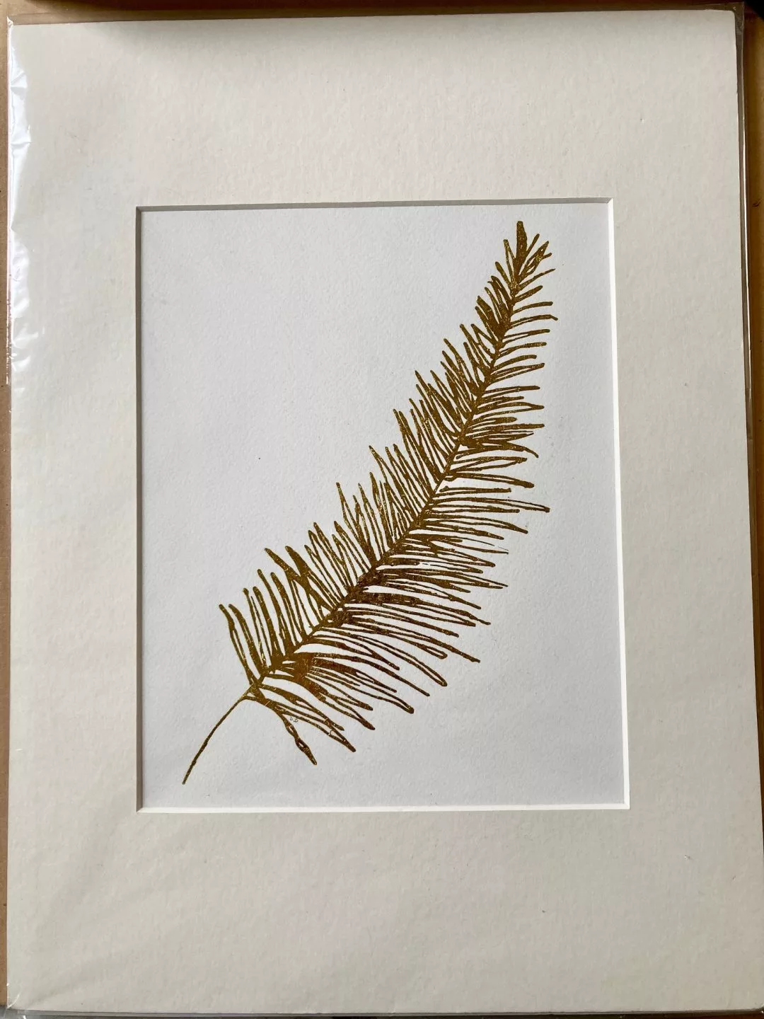 A framed artwork mounted on white mat board showing a single botanical frond printed in brown‑gold ink. The frond is slender and slightly curved, with many fine, narrow leaflets extending from a central stem, resembling a fern or conifer branch. It is positioned diagonally across a plain white background. The paper has a soft, textured surface, and the overall composition is minimal, focusing attention on the delicate linear details of the plant form.