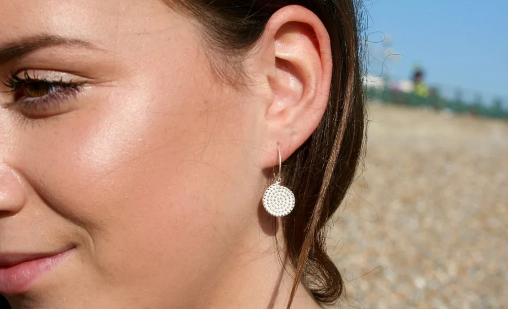 A close-up view of one silver drop earring being worn on an ear. The earring has a thin curved hook and a round disc-shaped charm with a textured pattern made of small raised bumps in concentric circles. The background is outdoors, with a blurred area showing pebbles and a blue sky.