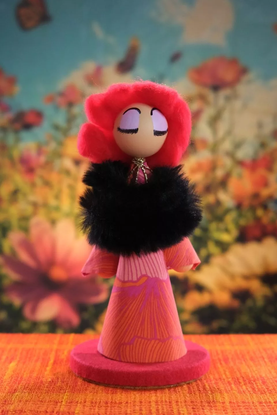 A small figurine displayed against a blurred floral background. The figure has a rounded head with painted closed eyes and long eyelashes. The hair is bright red and voluminous, styled in soft rounded shapes. The body is cone‑shaped, covered in patterned pink and orange fabric. Around the neck and shoulders is a thick black fluffy collar. The figure stands on a circular red base placed on an orange textured surface.