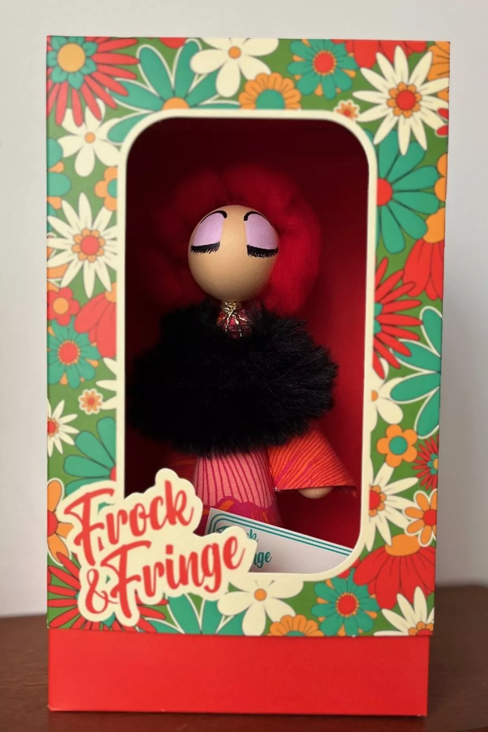 A figurine is packaged inside a rectangular display box. The box has a floral pattern in red, green, yellow, and white around the edges, with a clear window on the front showing the figure inside. The figurine has a rounded head with closed painted eyes, bright red hair, a black fluffy collar, and a pink and orange patterned cone‑shaped body. The bottom of the box is solid red, and a label reading “Frock & Fringe” is printed on the front in stylised script.