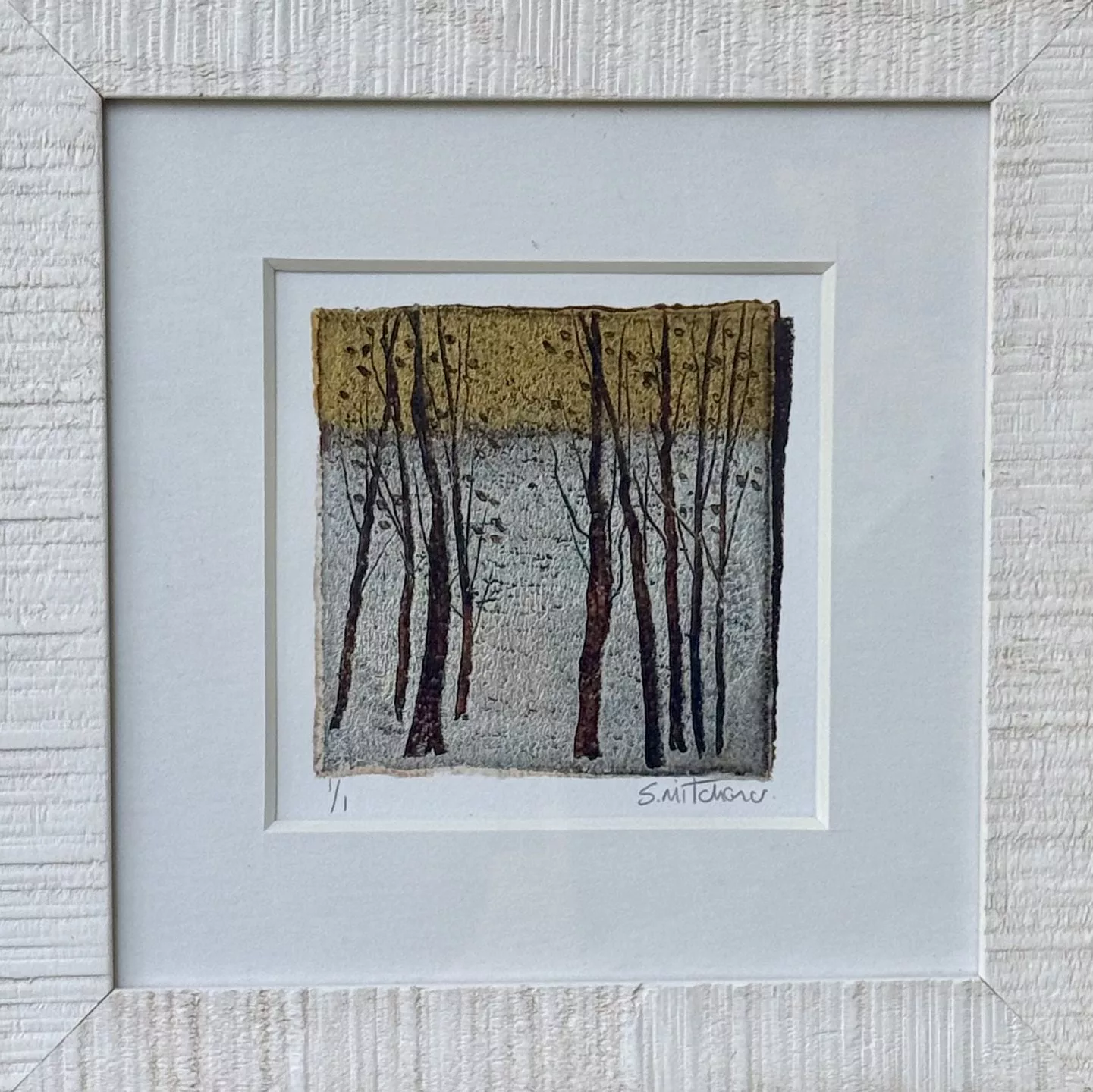 A small square artwork displayed inside a wide white textured frame. The artwork features a group of tall, thin tree trunks with bare branches, shown in dark brown and black tones. The trees stand against a two‑part background: the upper half is a muted golden‑yellow, and the lower half is a pale grey. The surface of the artwork has a textured, handmade appearance with rough, uneven edges. At the bottom of the artwork are handwritten markings, including “1/1” on the left and a signature on the right.