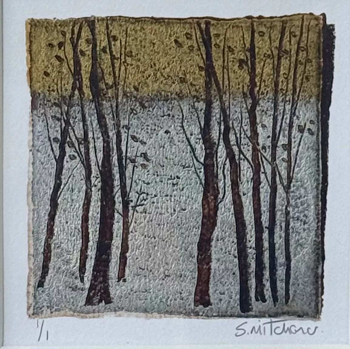 A small square artwork displayed inside a wide white textured frame. The artwork features a group of tall, thin tree trunks with bare branches, shown in dark brown and black tones. The trees stand against a two‑part background: the upper half is a muted golden‑yellow, and the lower half is a pale grey. The surface of the artwork has a textured, handmade appearance with rough, uneven edges. At the bottom of the artwork are handwritten markings, including “1/1” on the left and a signature on the right.