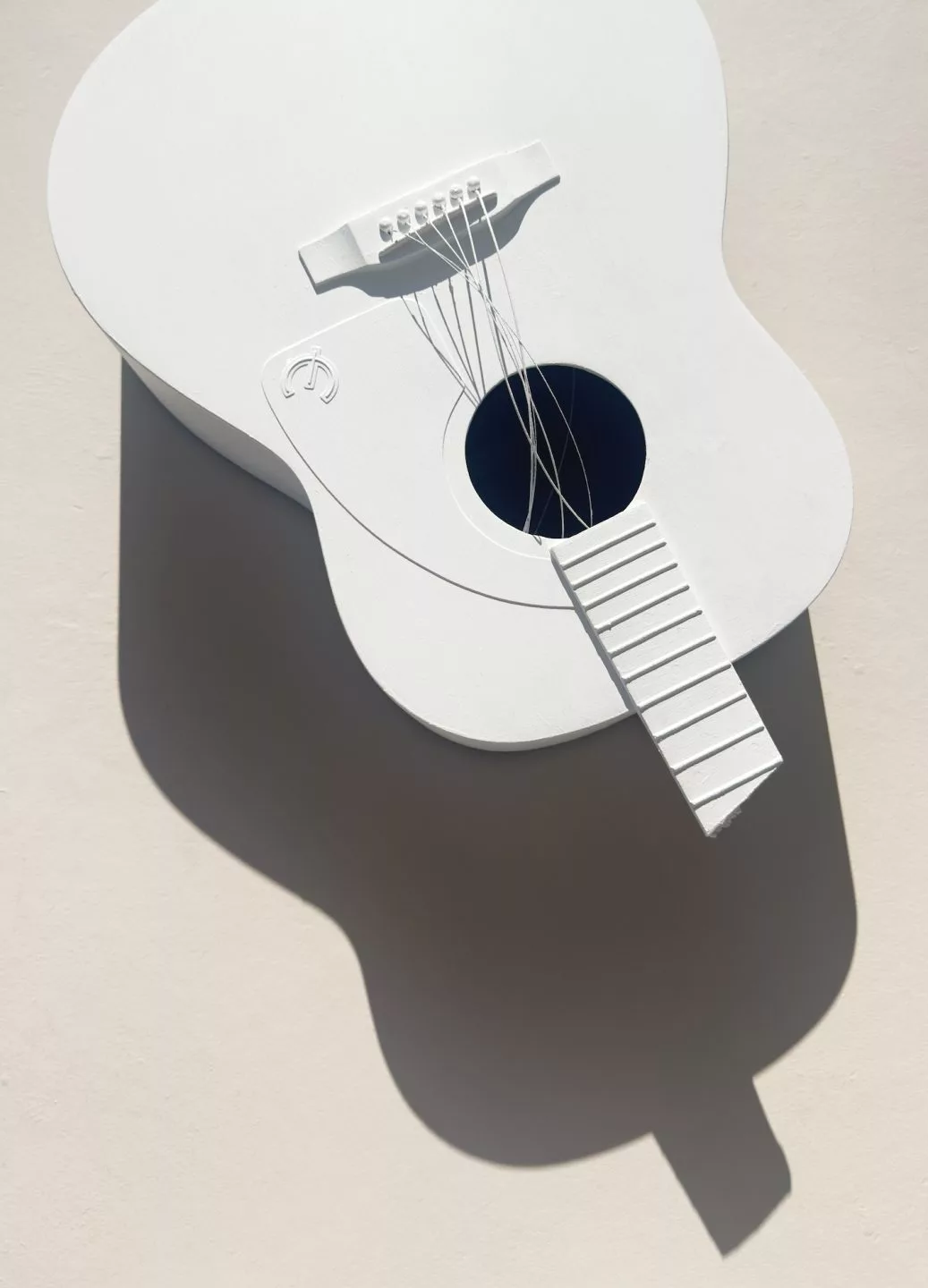 A white sculptural object in the shape of an acoustic guitar is shown lying on a light surface. The guitar has simplified, stylised details, including a flat body, a round sound hole, raised bridge, and a fretboard without marked frets. Thin white wires represent the strings, some loosely curved and not fully attached. The entire guitar is monochromatic white, creating strong shadows cast to the left due to bright directional lighting. The shadow forms an elongated, soft‑edged silhouette of the guitar’s body and neck on the background.