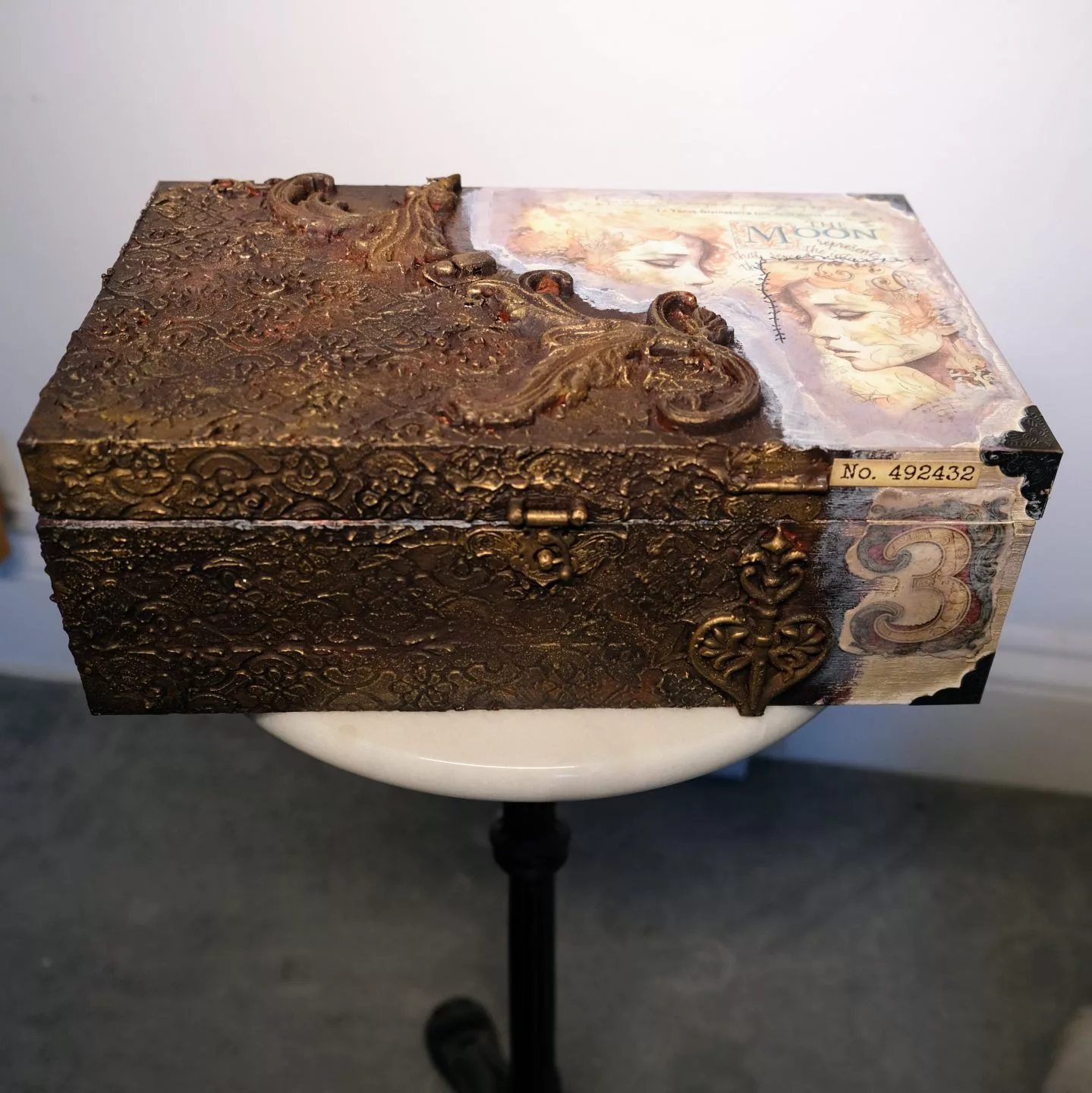 A decorated rectangular box with an ornate bronze‑textured surface, featuring raised patterns and small sculptural details. One side shows a collage‑style image with a classical face and printed numbers. The box rests on a white stool against a neutral background.