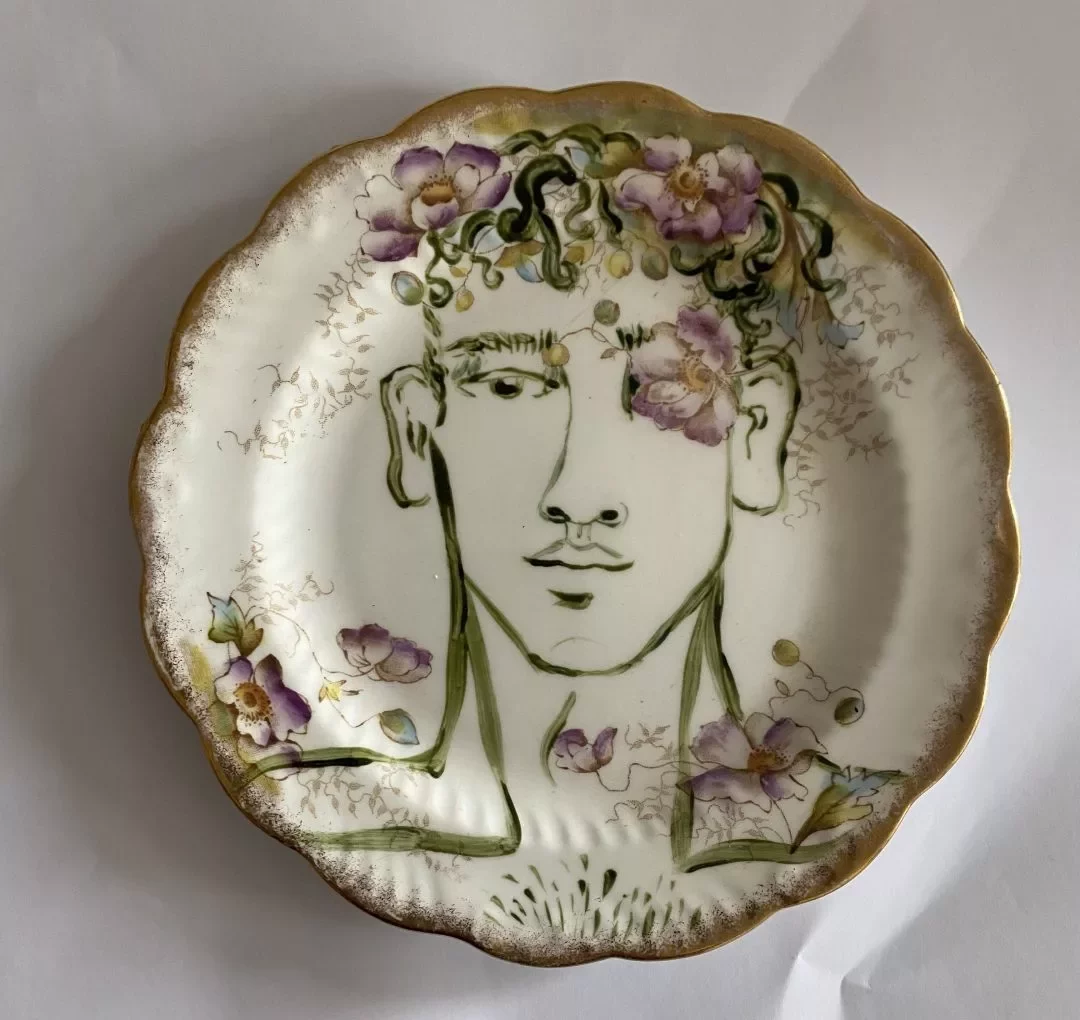A decorative ceramic plate with a scalloped, gold‑rimmed edge is shown against a plain white background. The center of the plate features a line‑drawn portrait of a person from the shoulders up. The portrait is drawn in dark green lines, showing the head, neck, and upper torso. Surrounding the head and hair are hand‑painted floral designs in soft purples, pinks, and greens, some appearing to blend into the hair. Additional small floral elements are scattered around the lower part of the portrait. The plate’s surface has a vintage, slightly ornate style, with gold detailing around the rim.