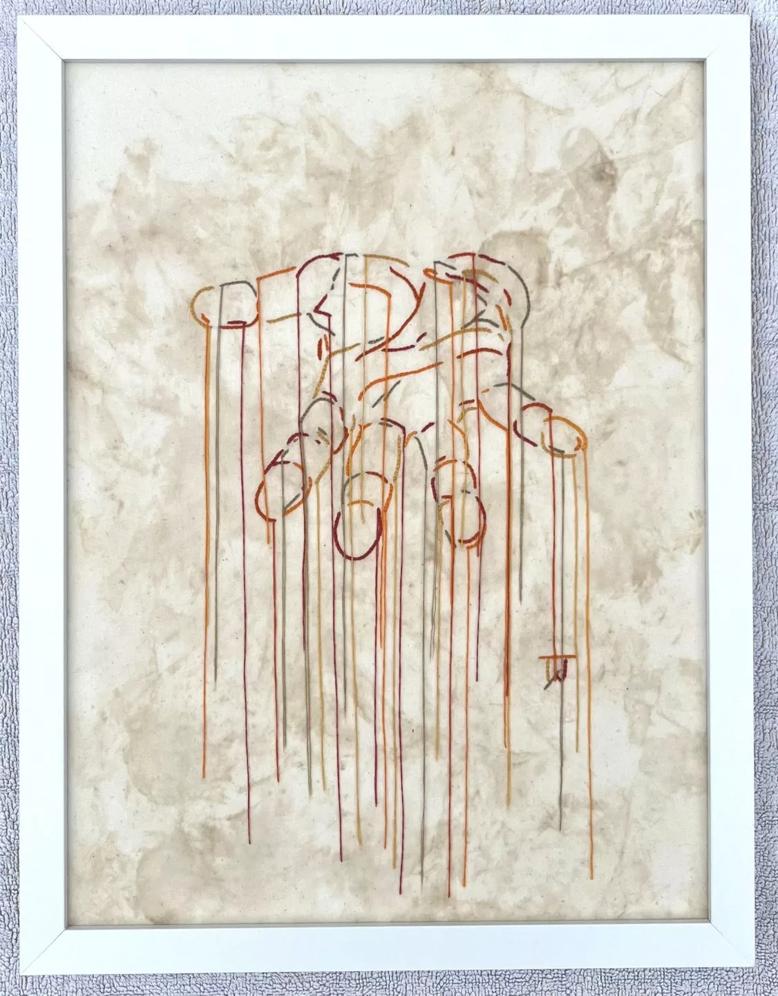 A minimalist artwork on a plain, off‑white canvas showing the outline of a open human palm created entirely from long, colorful embroidery threads. Multiple strands of thread in shades of blue, red, yellow, and orange trail downward from each point of the palm, creating long vertical lines that hang loosely toward the bottom of the canvas. The style is delicate and airy, giving the impression of motion and suspension. The image is framed in a simple white frame.