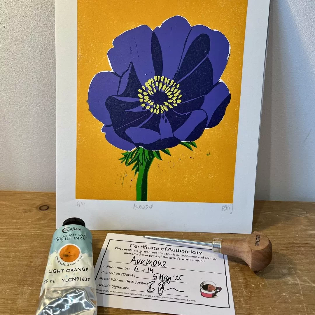 A framed print of a large purple anemone flower with layered petals and a yellow center is propped upright against a wall on a wooden surface. The background of the print is a solid bright orange. Below the print lies a certificate of authenticity, a metal tube of light‑orange printing ink, and a wooden‑handled roller tool.