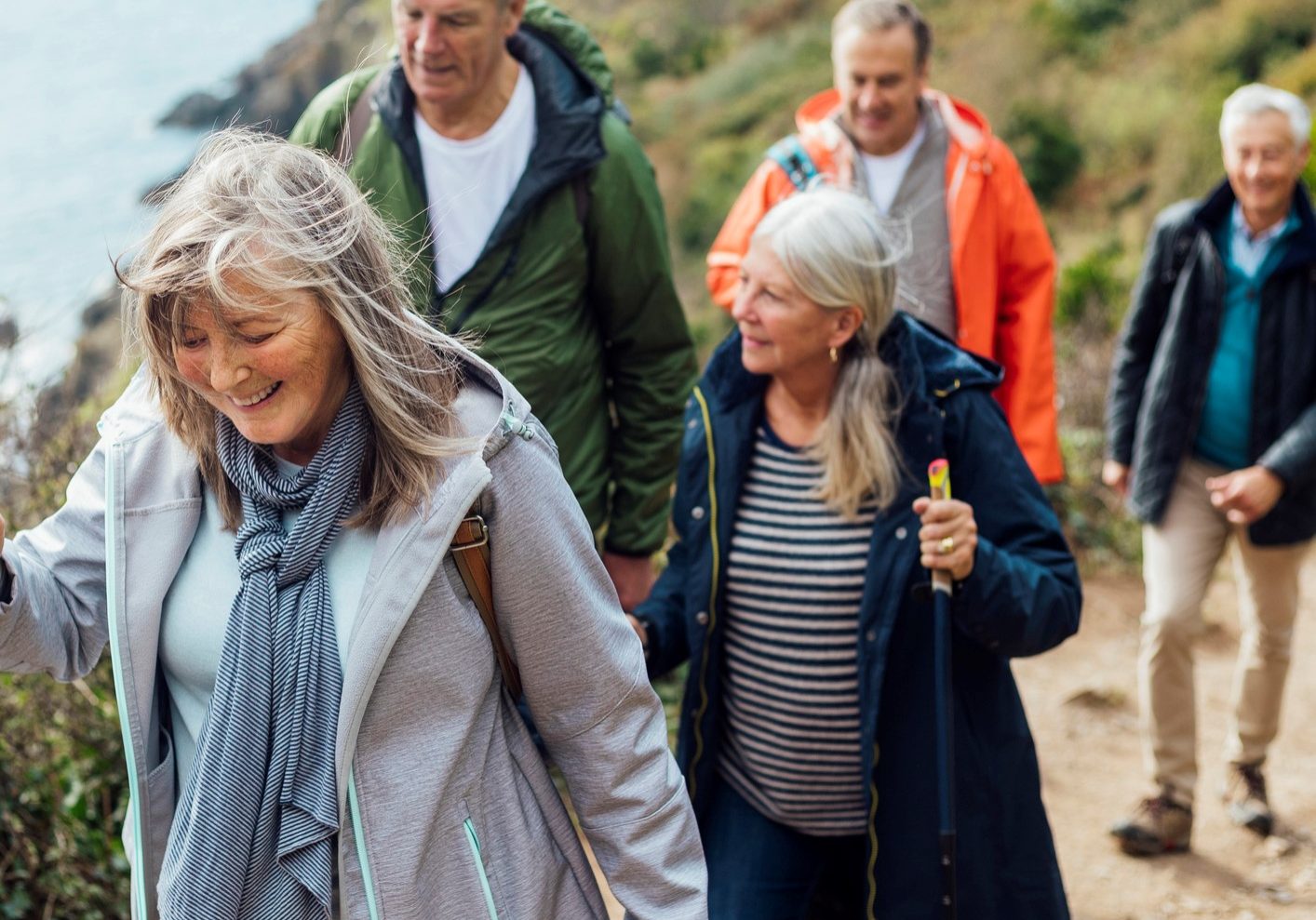 A group of male and female friends walking up a hill on a coastal path along a cliff edge overlooking the sea in Cornwall. The main focus is one woman wearing a jacket and scarf, she is smiling while looking down.