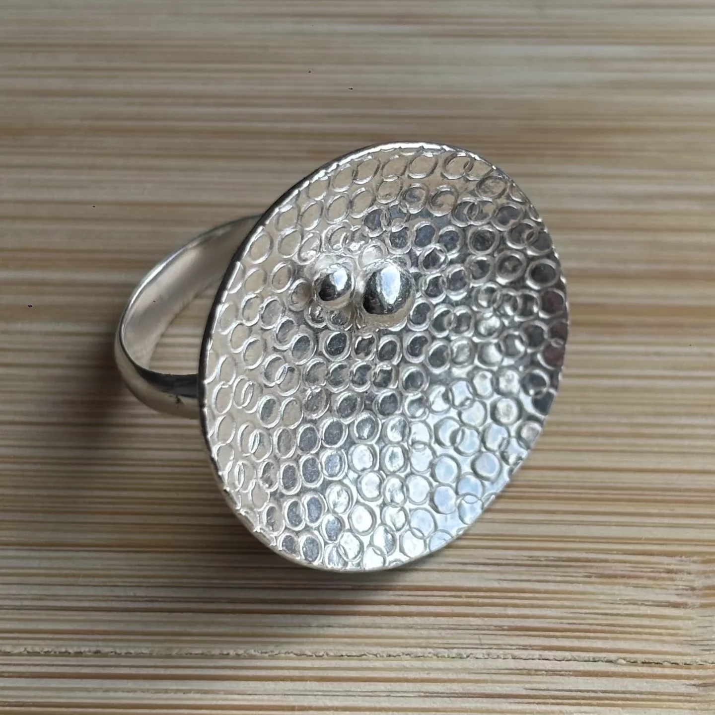 A silver ring displayed on a light wooden surface with fine horizontal grain lines. The ring features a large, flat, oval‑shaped top with a hammered texture made up of small, evenly spaced circular impressions. Near the center of the oval are two raised polished silver dots, one slightly larger than the other. The ring band is smooth and rounded, attached to the back of the oval top. The metal surface reflects light softly, highlighting the textured pattern.