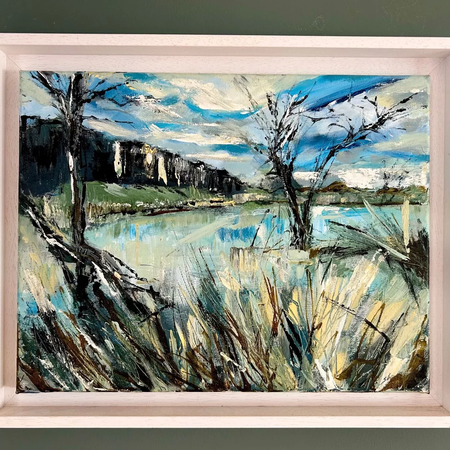 A framed landscape painting showing a riverside or lakeside scene. In the foreground, tall grasses and reeds in shades of beige, green, and brown stretch upward with bold, textured brushstrokes. Behind them, calm reflective water occupies the center of the painting, capturing patches of light blue and soft yellow highlights. On either side of the water, two leafless trees with dark, expressive branches stand partly submerged. A long, dark hillside runs across the left side of the background, contrasting with a bright sky filled with streaks of blue, white, and grey cloud patterns. The painting is framed in a light wooden frame and is shown hanging against a muted green wall.