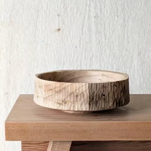 A handcrafted wooden bowl sits on a wooden surface. The bowl has a rough, textured exterior with visible carved grooves and tool marks, giving it a rustic, handmade feel. Its interior is smooth and polished, showing the natural grain of the wood. Behind it is a plain, light‑coloured wall that keeps the focus on the bowl’s craftsmanship and organic texture.