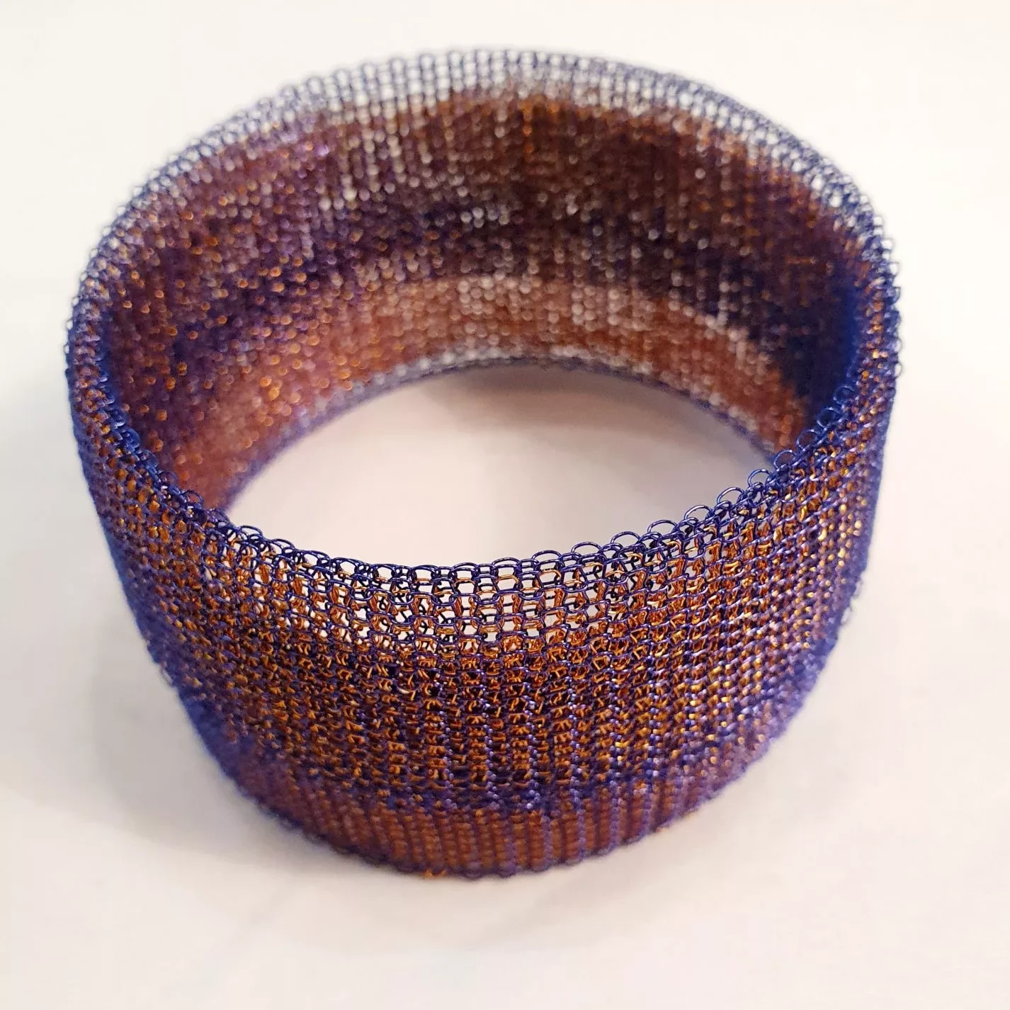 A wide bangle made from fine knitted wire mesh. The material is semi‑transparent, allowing light to pass through the open weave. The bangle features horizontal bands of color, shifting from deep purple to coppery brown tones. The structure appears lightweight and flexible, with a uniform cylindrical shape and evenly knitted texture. It is displayed on a plain white surface.