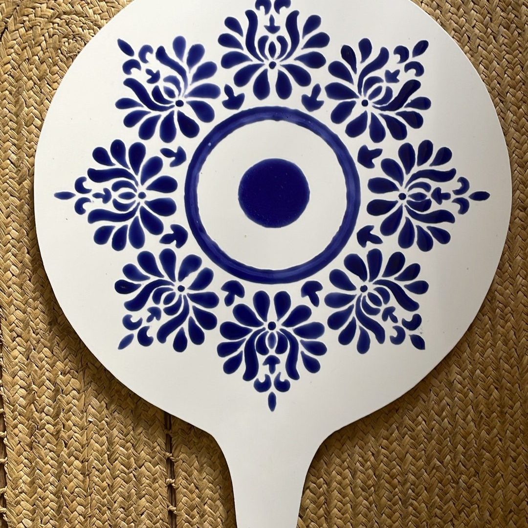 This is a white ceramic cheeseboard with a round top and small handle. It features an elegant blue and white design reminiscent of traditional porcelain patterns. The central motif has concentric circles with a solid blue dot in the middle, surrounded by ornate floral patterns arranged in a symmetrical medallion design. The board is displayed on a woven natural fiber mat or placemat with a braided texture.