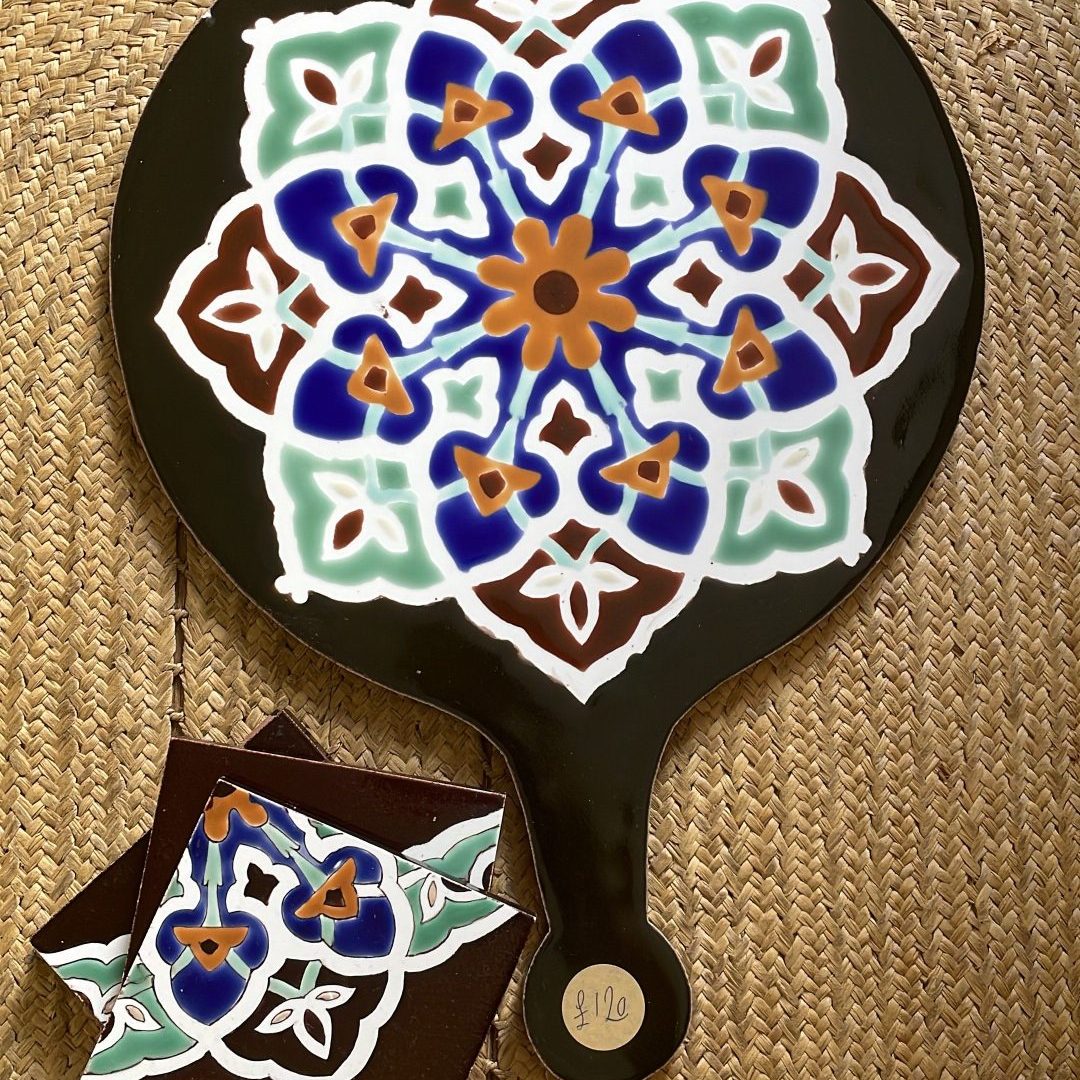 This ceramic cheeseboard has a black background with a colorful mandala-like pattern. The intricate design features a symmetrical arrangement of blue, mint green, white, and orange/brown elements forming a floral star pattern. The handle is visible at the bottom, and there appears to be a small matching coaster or sample piece with a similar pattern placed beside it. Like the first image, it's displayed on the same type of woven natural fiber mat.
