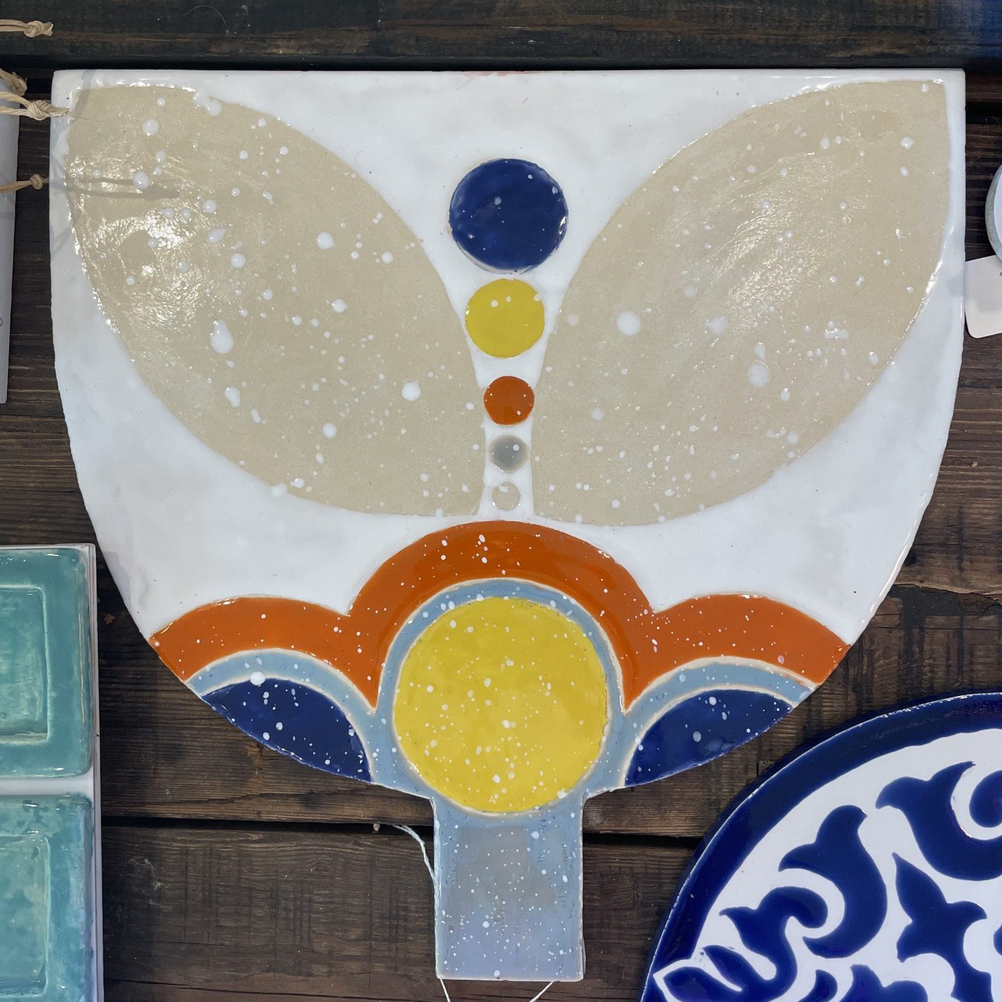 This artistic ceramic serving board has a white background with a stylized butterfly or angel-like design. The "wings" are a beige/cream color with white speckles, while the central "body" features a vertical line of colorful dots (blue, yellow, orange) and a decorative base with curved elements in blue, orange, and yellow. It has a small handle at the bottom and appears to be displayed on a wooden surface alongside other ceramic pieces, including what looks like turquoise tiles and a blue and white plate with a floral pattern.