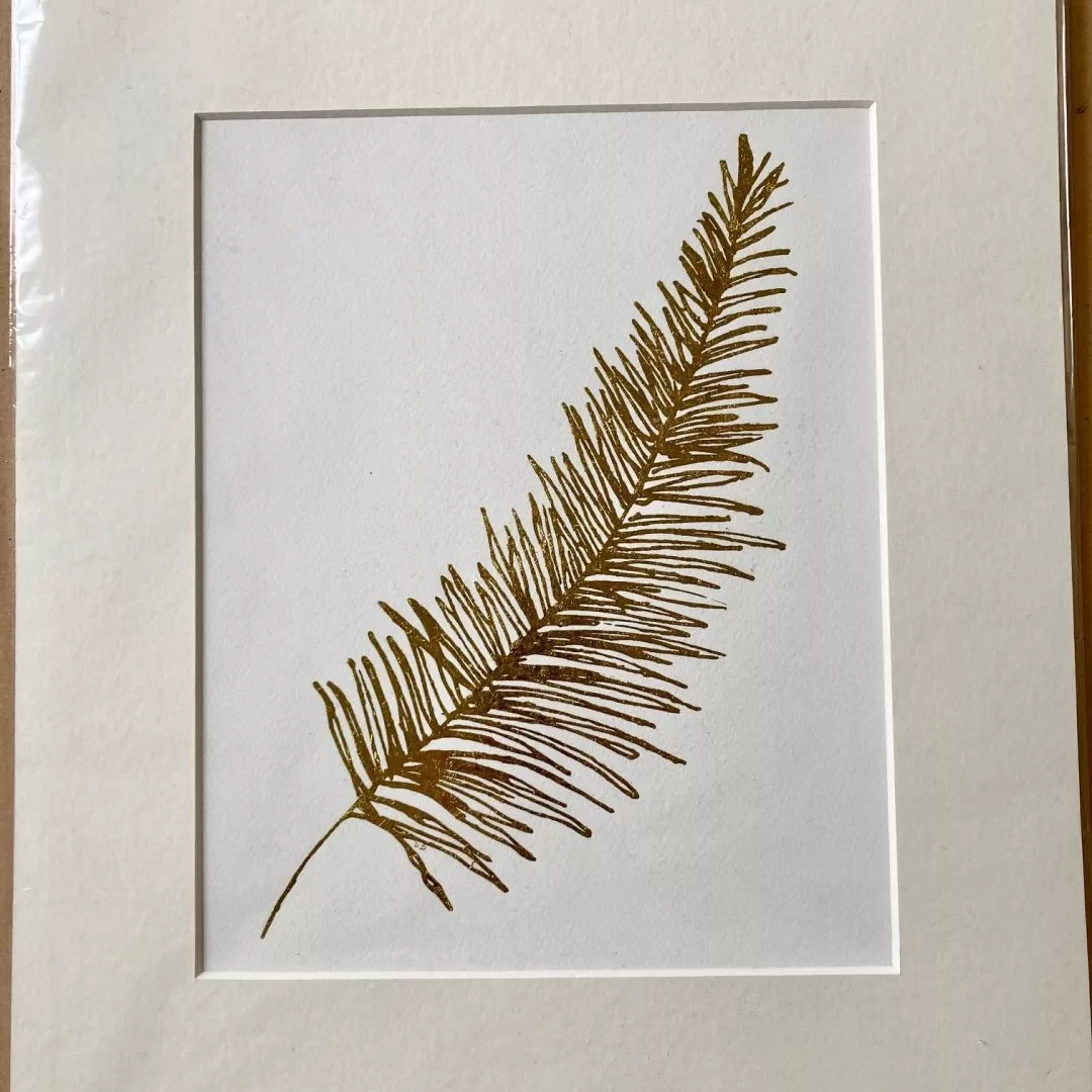 A framed artwork mounted on white mat board showing a single botanical frond printed in brown‑gold ink. The frond is slender and slightly curved, with many fine, narrow leaflets extending from a central stem, resembling a fern or conifer branch. It is positioned diagonally across a plain white background. The paper has a soft, textured surface, and the overall composition is minimal, focusing attention on the delicate linear details of the plant form.