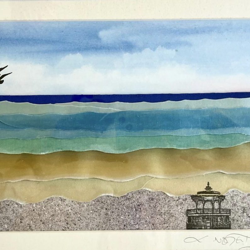 The image is a mixed media artwork titled Bandstand Blues, depicting a serene seascape. The scene is composed of layered, cut paper or translucent materials in soft gradients to represent the sea, sand, and sky. The sky transitions from light blue near the horizon to a deeper blue at the top, with soft white clouds. The sea is illustrated in gently undulating bands of turquoise, blue, and green, mimicking the movement of waves. Two black silhouettes of birds fly over the water in the distance, adding a sense of calm and motion. The foreground features layers of sandy tones leading to a speckled, grey shoreline. In the lower right corner, there is a fine black line drawing of a Victorian-style bandstand, adding a touch of architectural detail and nostalgia to the natural scene. The combination of collage and illustration creates a harmonious and peaceful coastal composition.