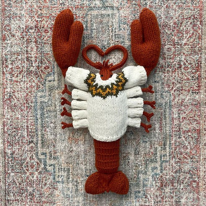 A hand‑knitted lobster plush lying on a patterned rug. The lobster has rust‑red claws, legs, and tail, with a cream‑coloured body and a decorative yellow, black, and brown motif around the top of the torso. The knitted texture is soft and detailed, giving the lobster a stylized, crafted appearance.