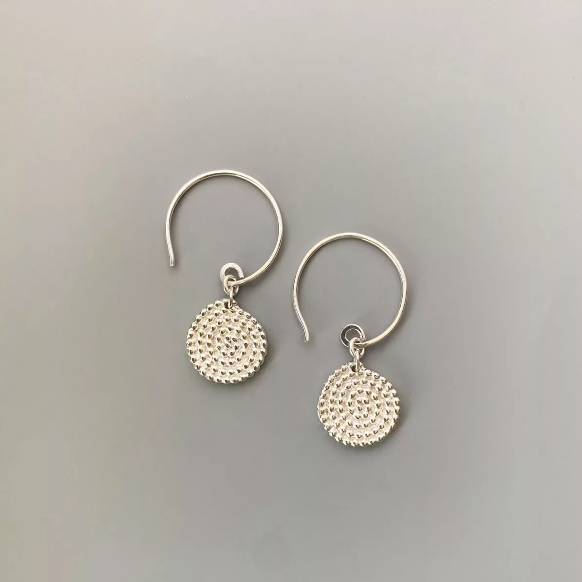 A pair of small silver drop earrings displayed on a smooth grey background. Each earring has a thin, circular hook and a round dangling charm. The charm is a flat disc covered in a raised, textured pattern made of tiny evenly spaced bumps arranged in concentric circles. The metal has a polished, reflective finish.