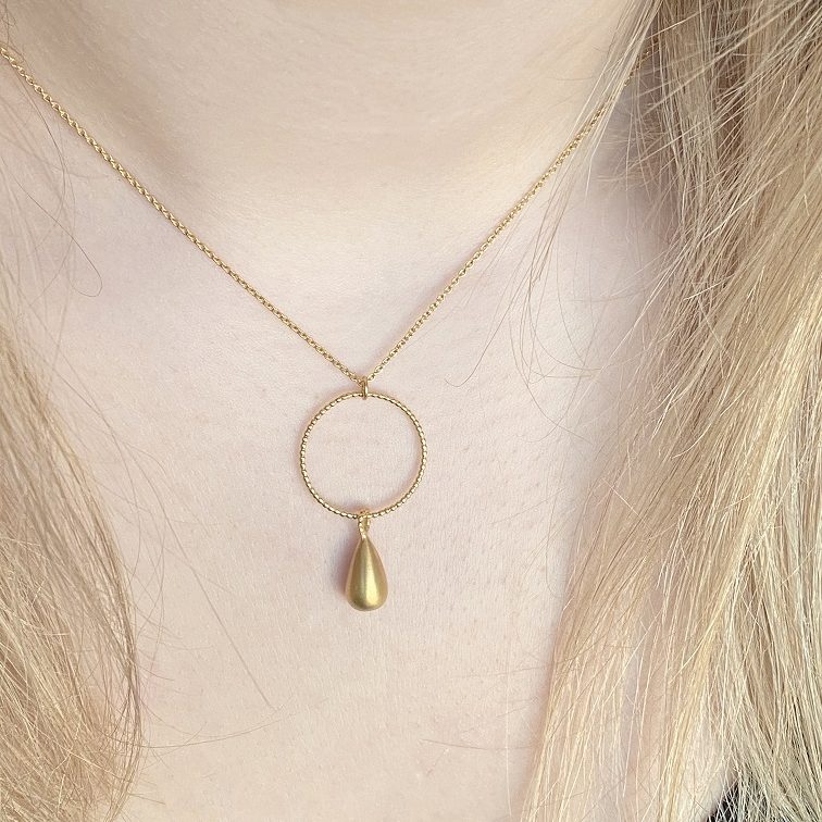A solid matte teardrop pendant gracefully suspended from a finely detailed, textured halo. This delicate yet striking design is crafted in warm-toned metal, offering a minimalist yet sophisticated statement. Suspended from a fine chain, the necklace exudes timeless elegance, making it a versatile accessory for any occasion.