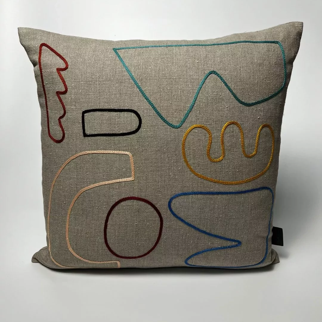 A square cushion with a light beige fabric cover shown against a plain white background. The front of the cushion features a series of abstract embroidered line shapes in various colors. These stitched lines include curved, looping, and angular forms in red, teal, yellow, black, peach, and blue. The shapes are arranged loosely across the surface without forming any recognizable image or text. The cushion appears soft and evenly filled, with slightly rounded corners and a small black fabric tag on the right edge.