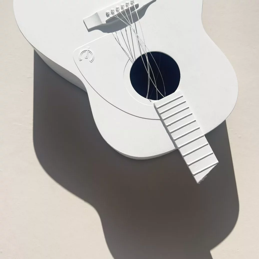 A white sculptural object in the shape of an acoustic guitar is shown lying on a light surface. The guitar has simplified, stylised details, including a flat body, a round sound hole, raised bridge, and a fretboard without marked frets. Thin white wires represent the strings, some loosely curved and not fully attached. The entire guitar is monochromatic white, creating strong shadows cast to the left due to bright directional lighting. The shadow forms an elongated, soft‑edged silhouette of the guitar’s body and neck on the background.