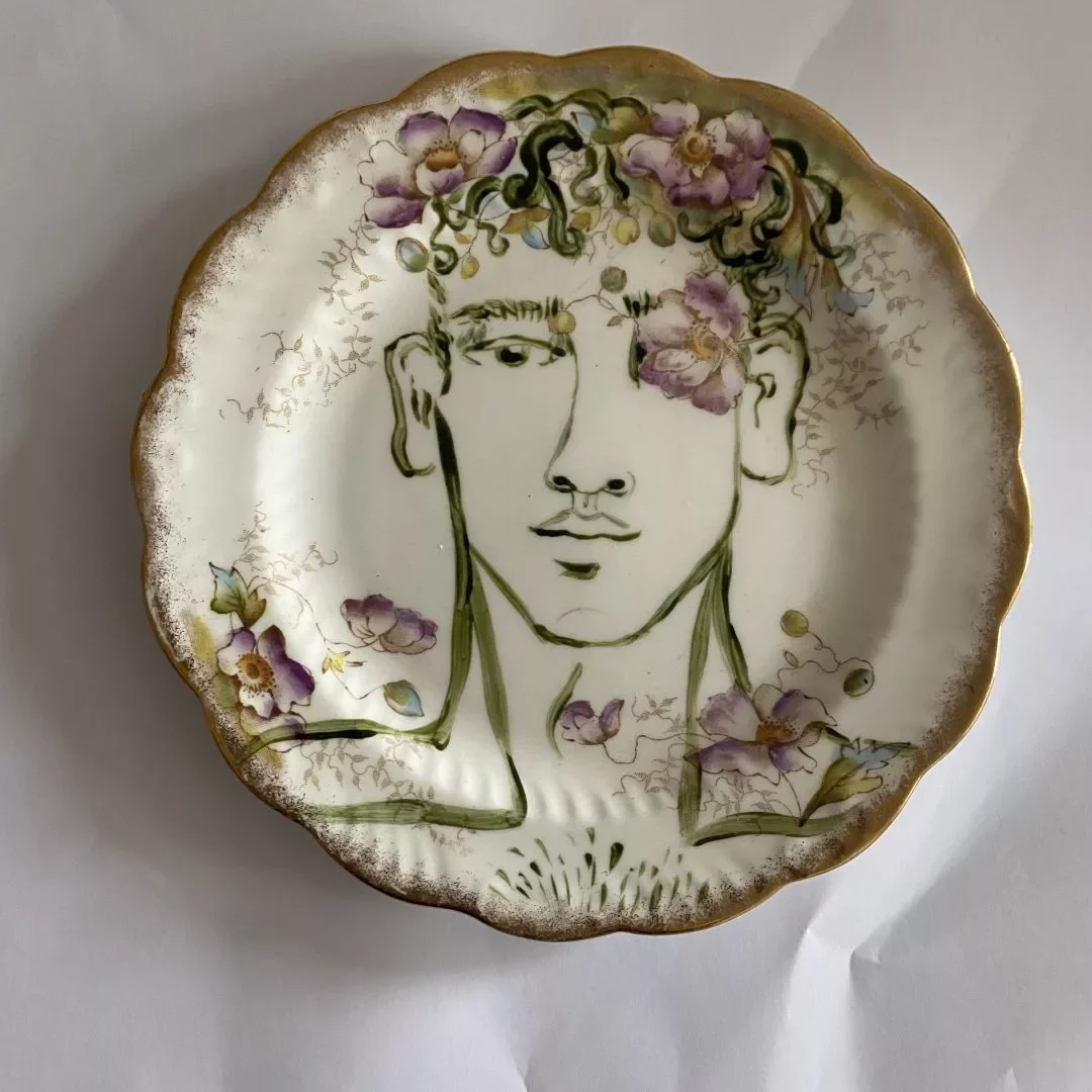 A decorative ceramic plate with a scalloped, gold‑rimmed edge is shown against a plain white background. The center of the plate features a line‑drawn portrait of a person from the shoulders up. The portrait is drawn in dark green lines, showing the head, neck, and upper torso. Surrounding the head and hair are hand‑painted floral designs in soft purples, pinks, and greens, some appearing to blend into the hair. Additional small floral elements are scattered around the lower part of the portrait. The plate’s surface has a vintage, slightly ornate style, with gold detailing around the rim.