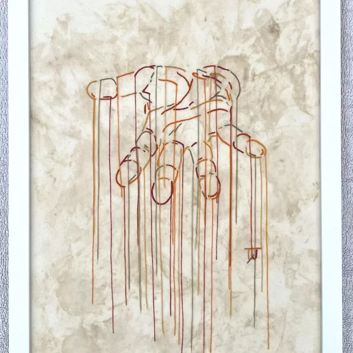 A minimalist artwork on a plain, off‑white canvas showing the outline of a open human palm created entirely from long, colorful embroidery threads. Multiple strands of thread in shades of blue, red, yellow, and orange trail downward from each point of the palm, creating long vertical lines that hang loosely toward the bottom of the canvas. The style is delicate and airy, giving the impression of motion and suspension. The image is framed in a simple white frame.